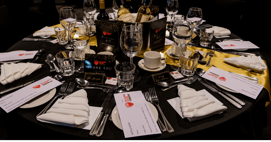 Image shows a circular dinner table, dressed in a black table cloth, with a full set of cutlery, plates, a special menu card and bottles of red and white wine.