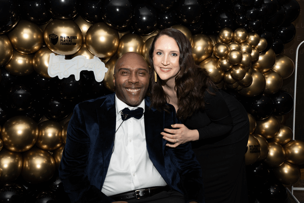 The image shows a man and woman standing together, smiling at the camera, dress in black tie attire.