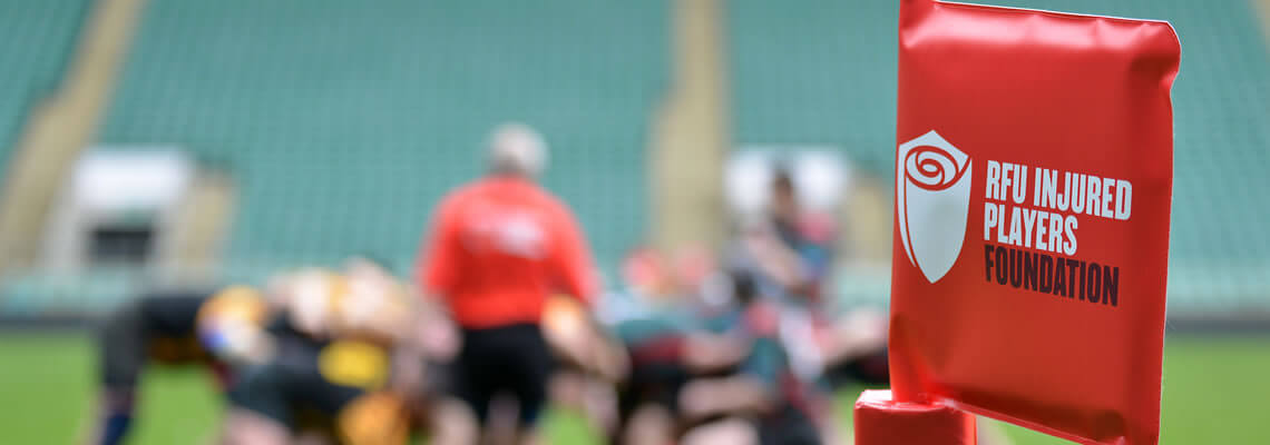Corner flag with IPF logo in focus in the foreground with a rugby scrum taking place out of focus in the background.